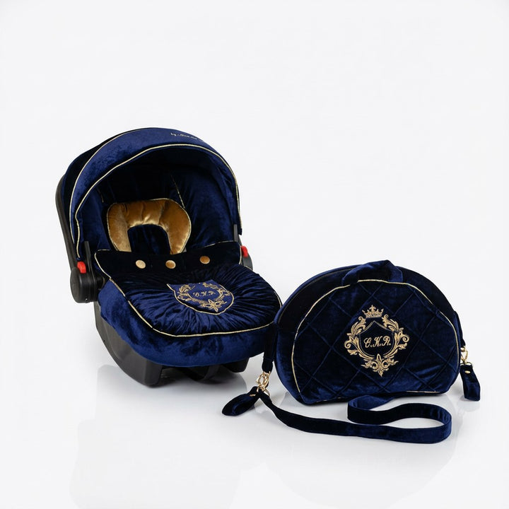 Dark navy blue velvet baby car seat set installed on a car seat, photographed on a white background. The hood has a structured rounded shape with light piping along the edge and a gold-colored inner accent. The seat cover features a central embroidered crest in gold thread. A matching navy velvet bag with gold monogram embroidery and crown detail is placed next to the car seat. All elements are shown from a front three-quarter angle.