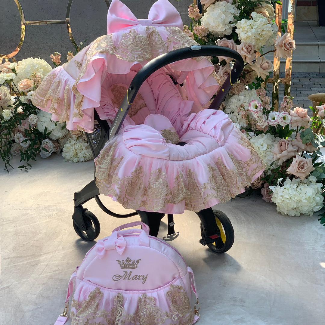 Pink luxury car seat cover set with gold lace ruffles and large bow on canopy, personalized diaper bag Mary with crown embroidery, floral backdrop.