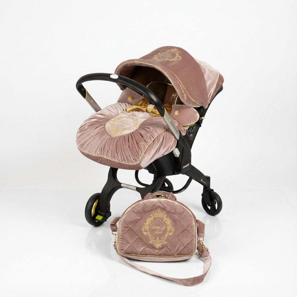Blush pink velvet Doona car seat cover set with gold trimming, gold embroidered crest, pleated footmuff and matching quilted bag on white background.