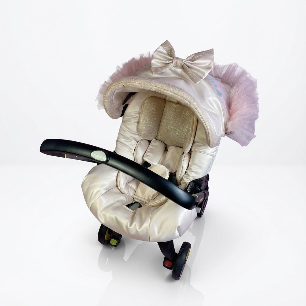 Front-view baby car seat cover installed on a car seat, photographed on a white background. The design features a soft ivory and champagne color palette with a padded canopy trimmed with delicate tulle ruffles. A satin bow decorates the top of the canopy, while the seat insert is quilted and softly padded for comfort.