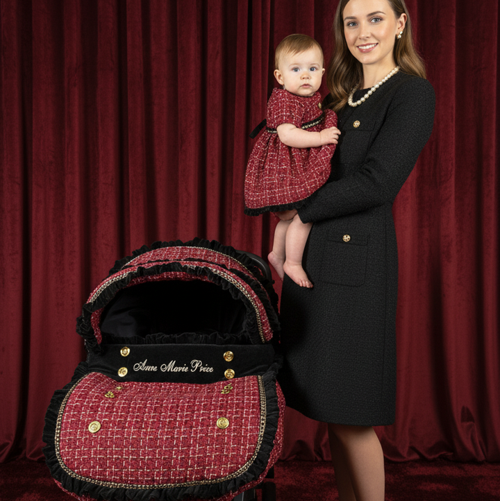 Woman holding a baby and standing next to a red and black baby carrier against a red curtain background.