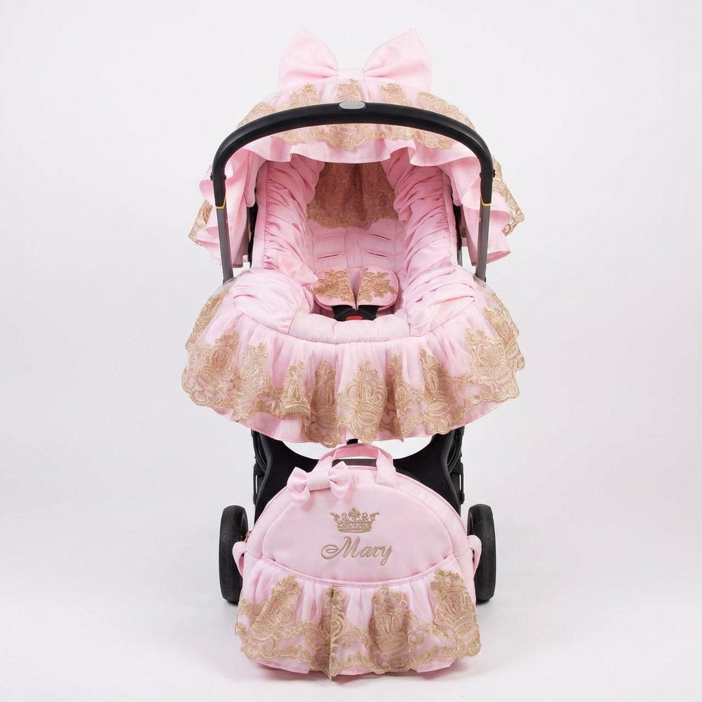 Pink satin infant car seat cover set with ruffled edges and gold lace trim, featuring a matching canopy with bow detail and a coordinating front bag embroidered with a crown and the name “Mary”, displayed on a black infant car seat frame against a white background.