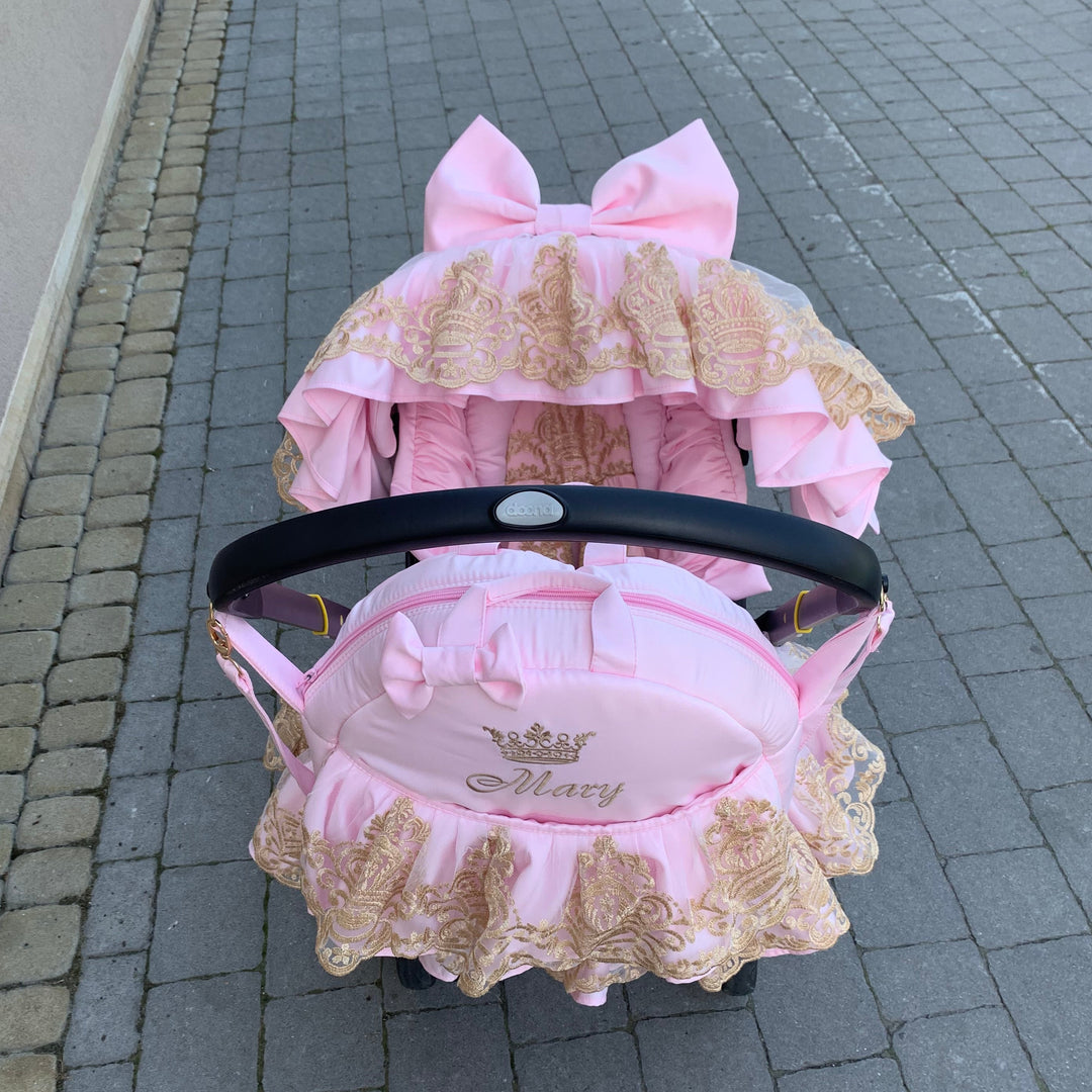 Pink luxury car seat cover set with gold lace ruffles and large bow canopy, personalized Mary embroidery, photographed outdoors on stone walkway.