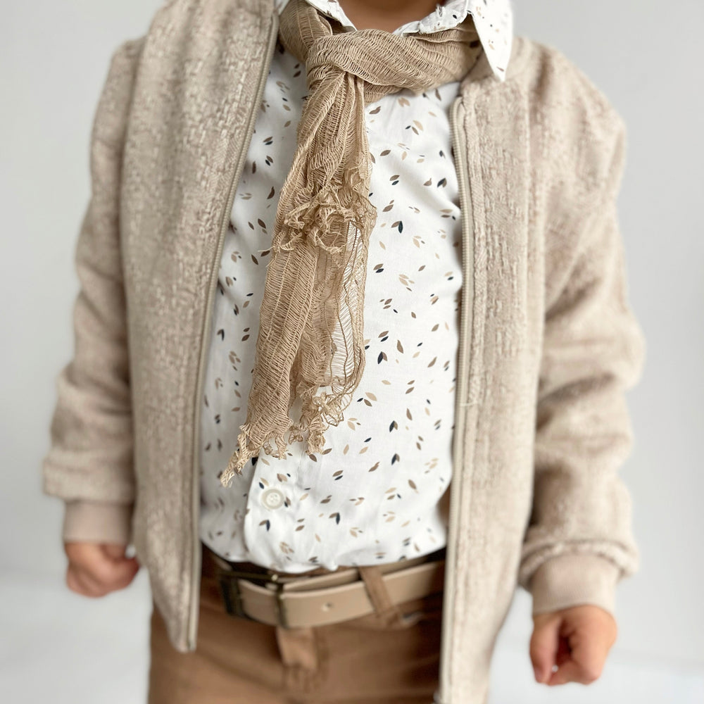 Person wearing a beige cardigan, patterned white shirt, and brown scarf on a plain background