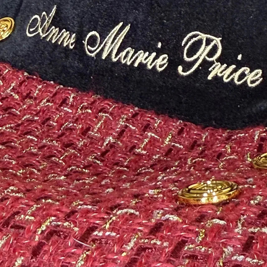 Close-up of a footmuff featuring white embroidered name “Anne Marie Price” on a black panel, with red tweed fabric woven with gold threads and decorative gold buttons.