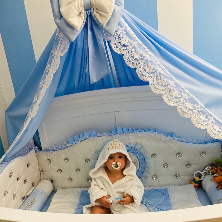 Child in a white robe with a hood sitting in a decorated crib with blue and white canopy.