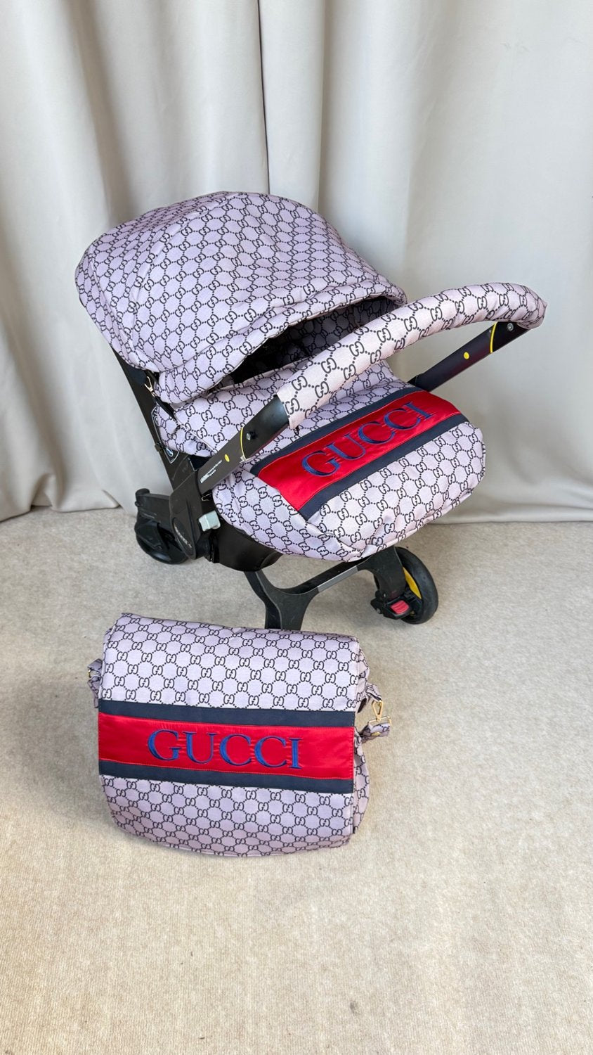 Doona car seat cover set with beige monogram pattern, red and navy stripe accent, and matching footmuff and bag.