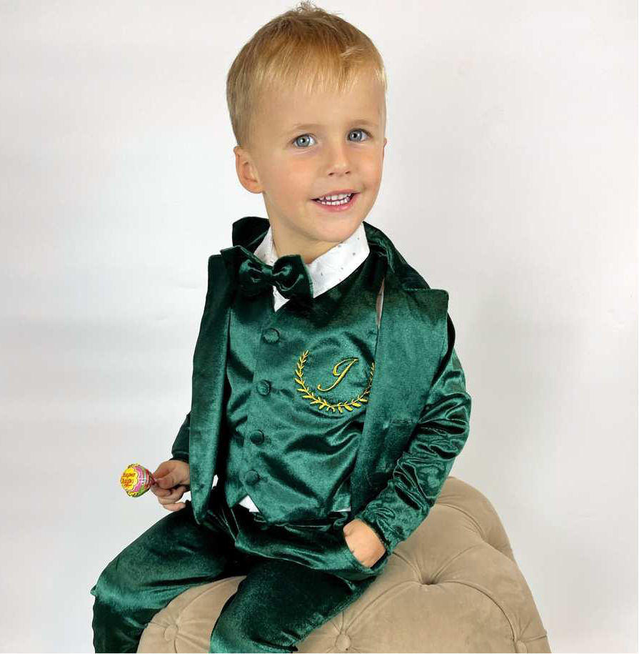 Young boy wearing a green velvet suit with a white shirt and bow tie, sitting on a beige chair against a white background.