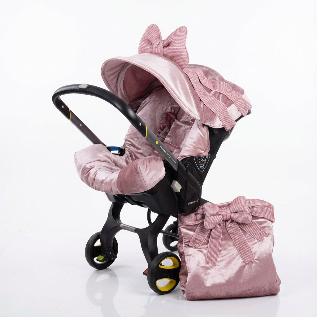 Blush pink velvet infant car seat cover with softly draped canopy and oversized decorative bow, paired with a matching velvet diaper bag, displayed on an infant car seat frame against a white background.