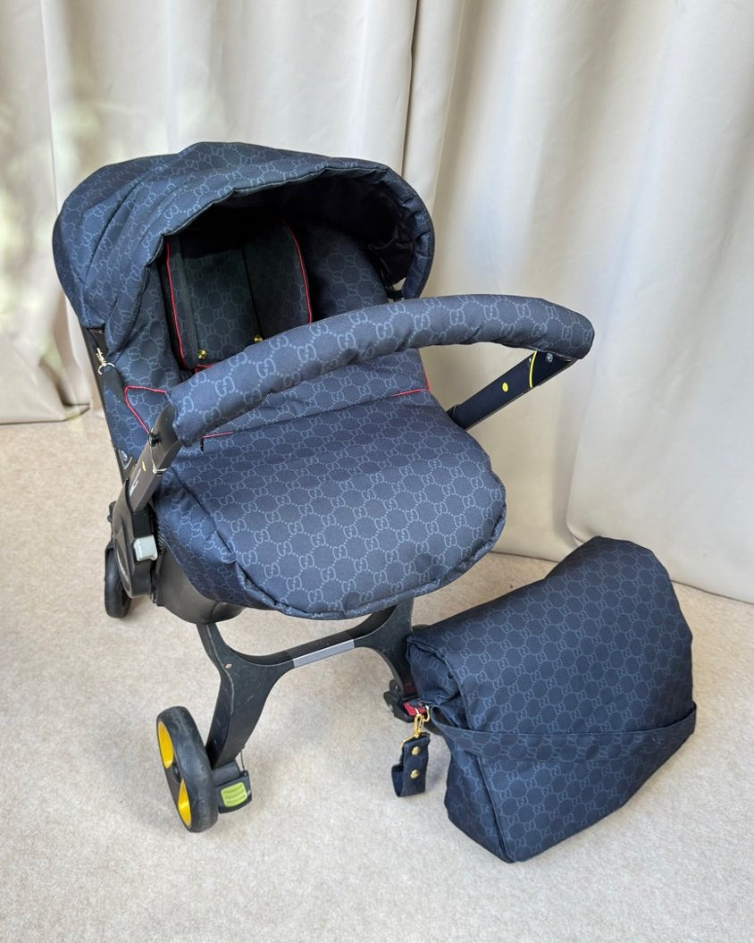 A navy blue baby stroller cover set with a matching footmuff and bassinet cover, featuring a repeating geometric monogram pattern, shown on a stroller with a beige carpeted floor and curtain background.