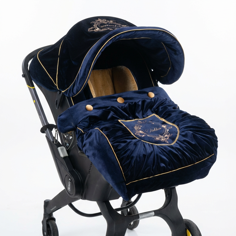 Royal blue velvet infant car seat cover with embroidered canopy, gold piping, decorative buttons, and a draped front panel with ornate crest embroidery, shown on a wheeled car seat frame against a white background.