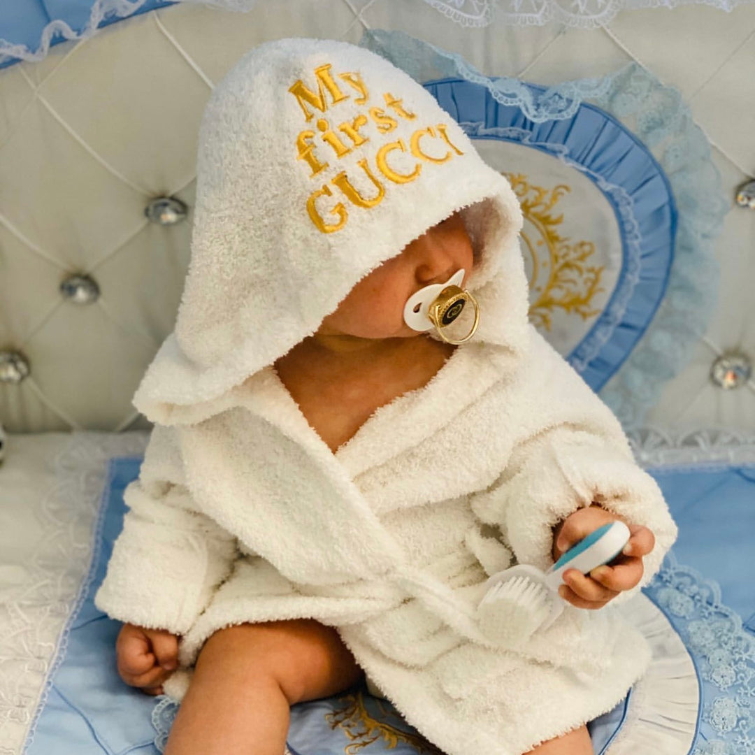 Baby wearing a white robe with 'My first Gucci' text on a decorative chair.