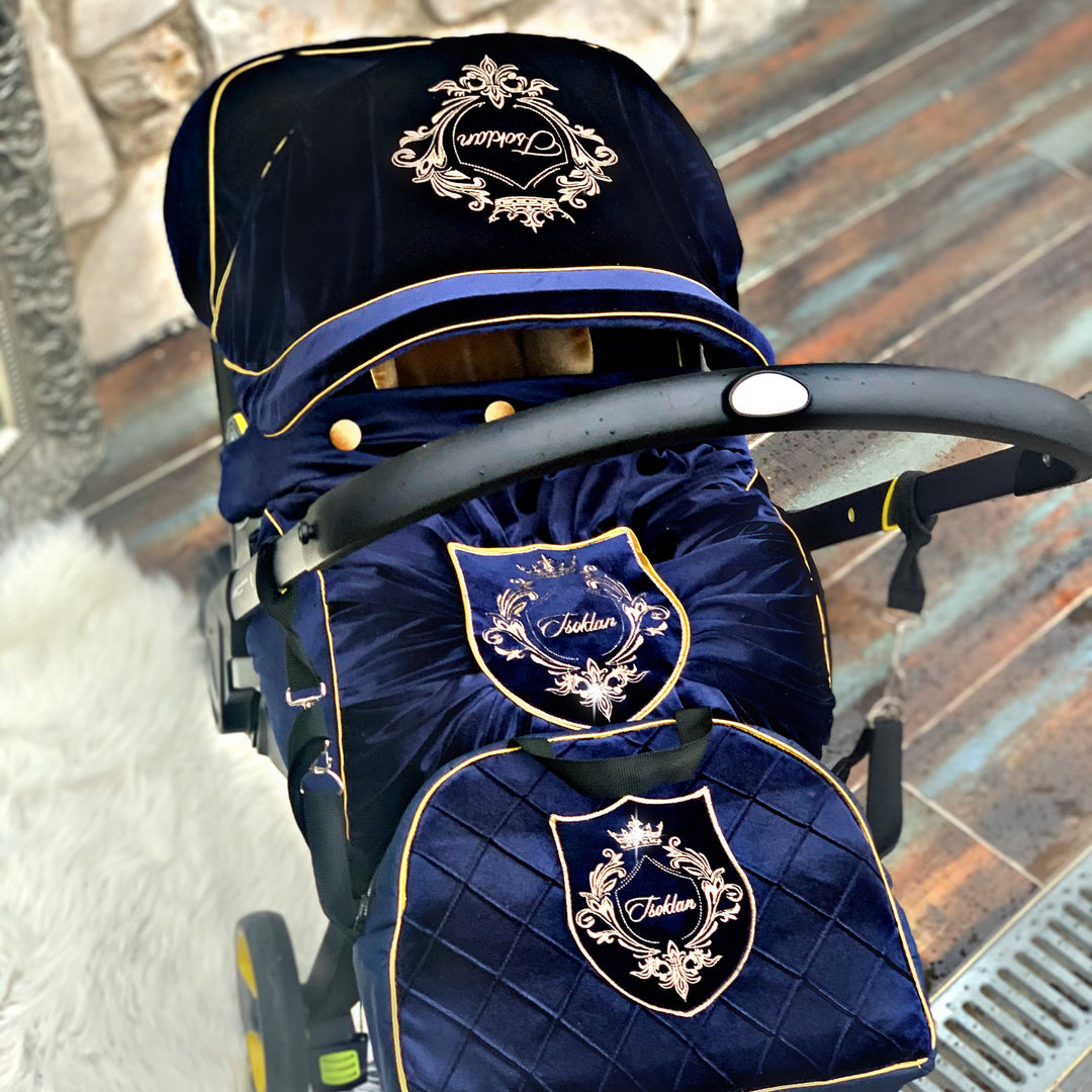 Top view of navy velvet Doona Plus car seat cover set with gold crest embroidery and matching quilted diaper bag on rustic stone wall and aged rust-effect tile background.