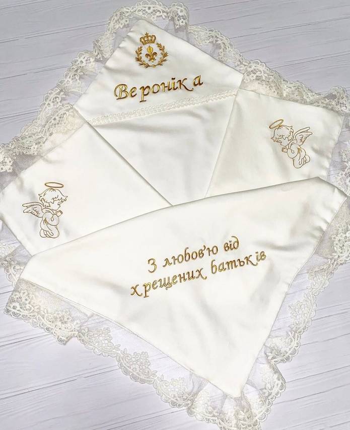 A personalized baptism blanket with ivory color, gold embroidery, and French lace detailing. It features custom text and a cross motif.