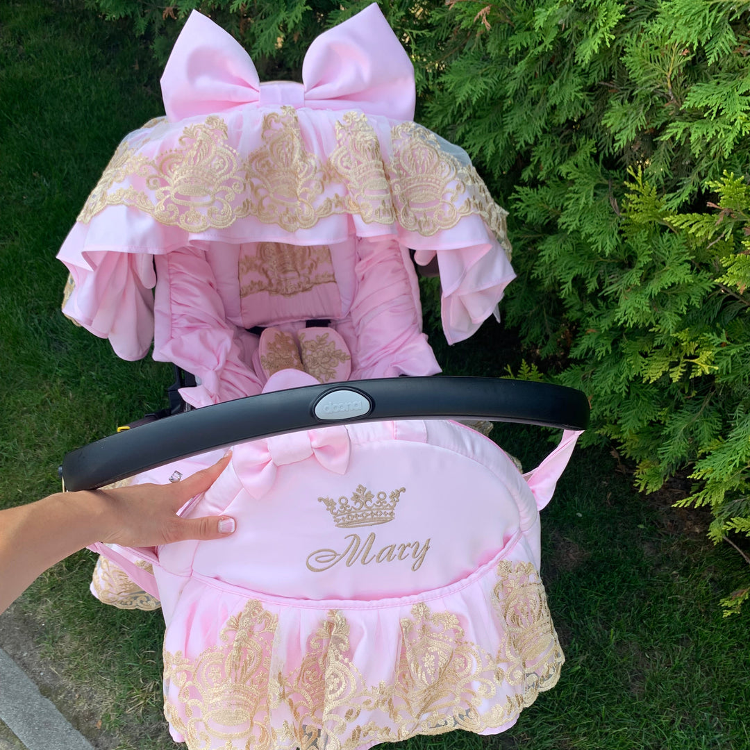 Luxury pink car seat cover set with gold lace ruffles and large bow canopy, personalized Mary embroidery, photographed outdoors against green hedge.