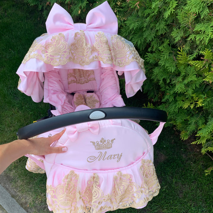 Luxury pink car seat cover set with gold lace ruffles and large bow canopy, personalized Mary embroidery, photographed outdoors against green hedge.
