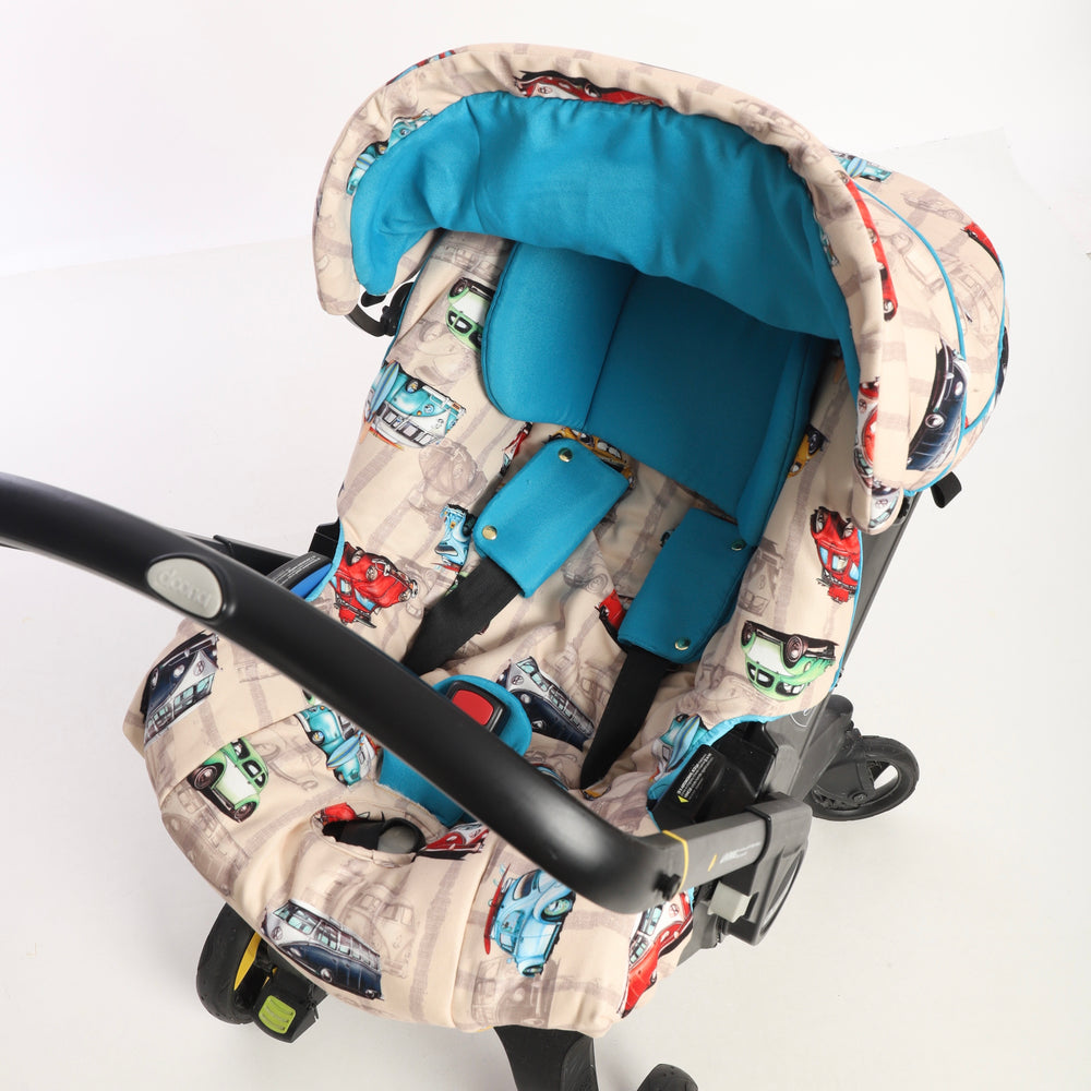 Front-facing baby car seat cover set installed on a car seat, featuring a retro vehicle print with classic Volkswagen Beetles and vintage minibuses in red, green, blue, and light blue on a beige background. The set includes a matching canopy and padded seat cover with turquoise inner padding and shoulder pads, displayed on a white background.