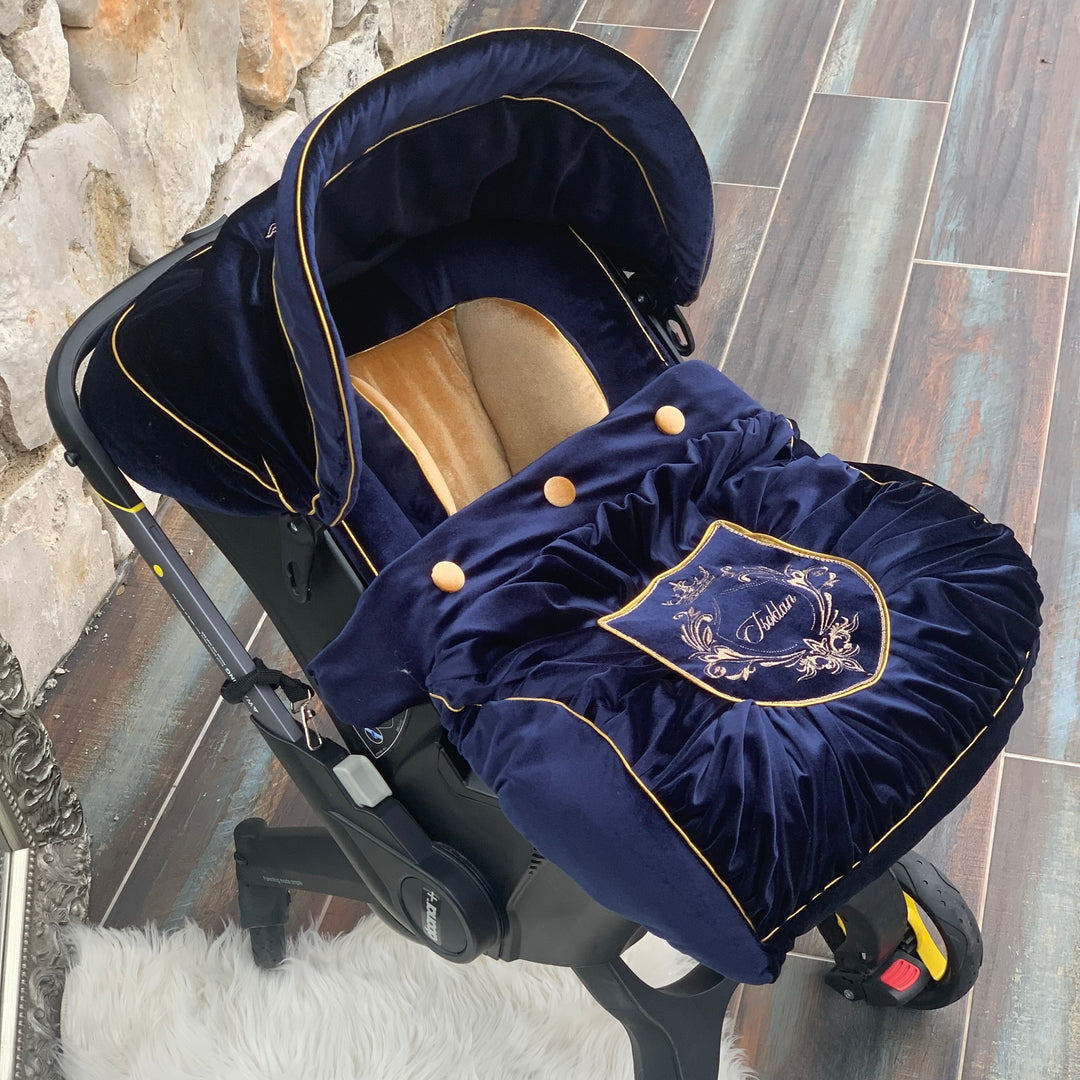 Royal Blue velvet Doona Plus car seat cover with gold trimming, gold buttons and embroidered crest, featuring warm gold insert, styled on rustic tile floor with stone wall background.