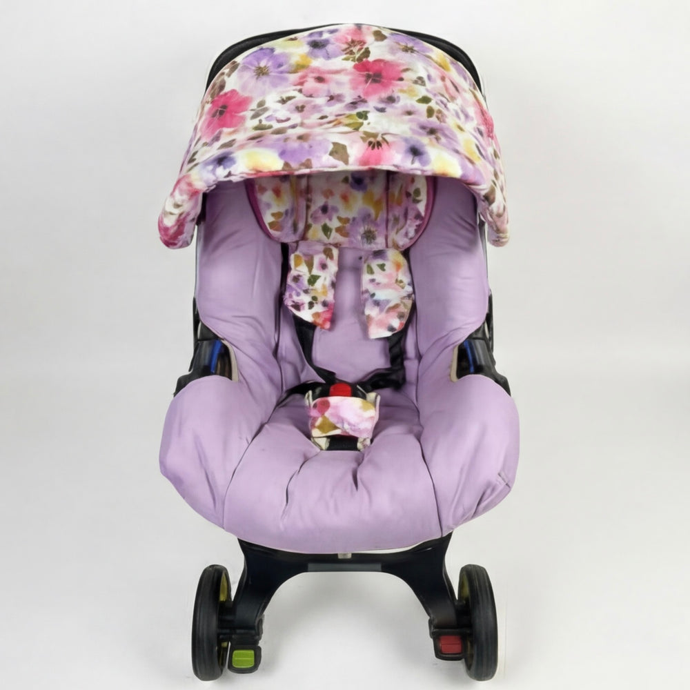 Front view of a baby car seat set installed on a car seat, featuring a soft lavender seat insert with matching padded sides, floral-patterned shoulder and crotch pads, and a pastel floral canopy in pink, lilac, yellow, and green tones. Shown on a clean white background.