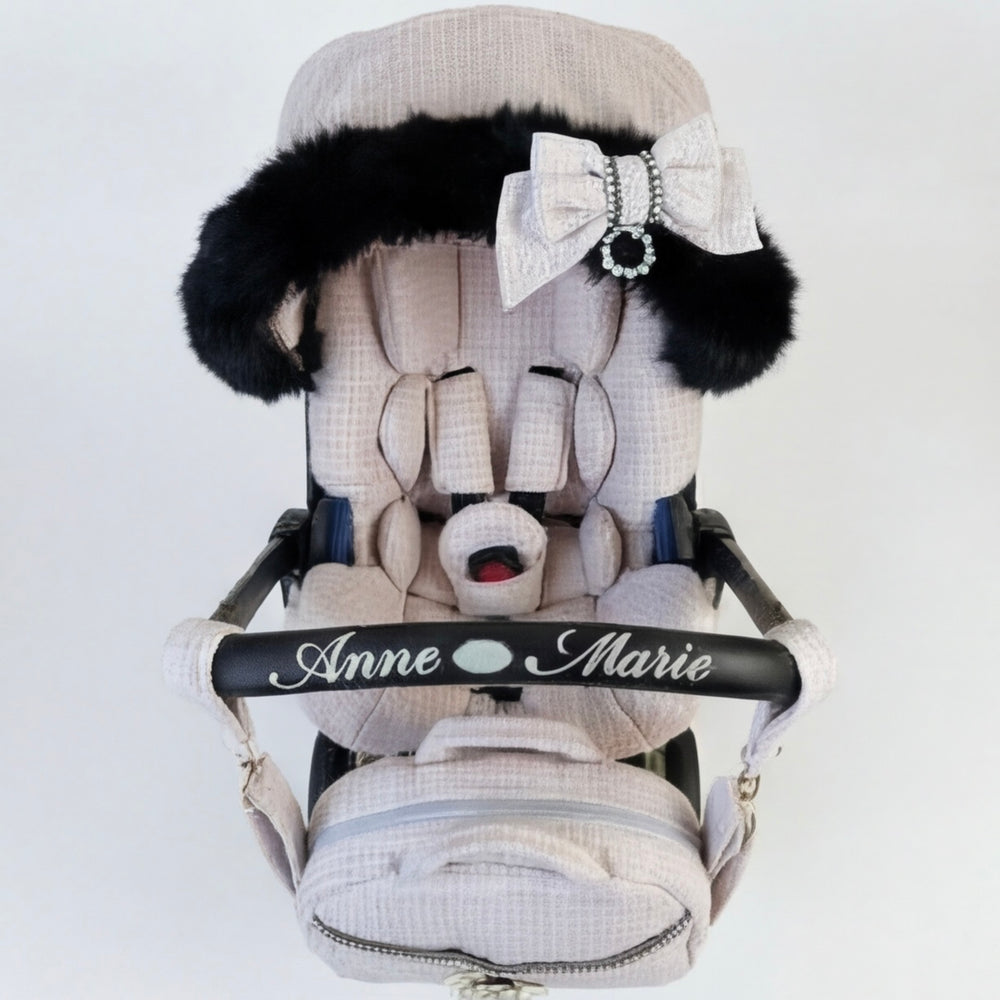 Front view of a baby car seat installed on a stroller frame, upholstered in light pink tweed fabric. The canopy is trimmed with real black fur and decorated with a light pink bow. The carry handle features embroidered text “Anne Marie.” A matching padded footmuff is attached. Displayed on a plain light background.