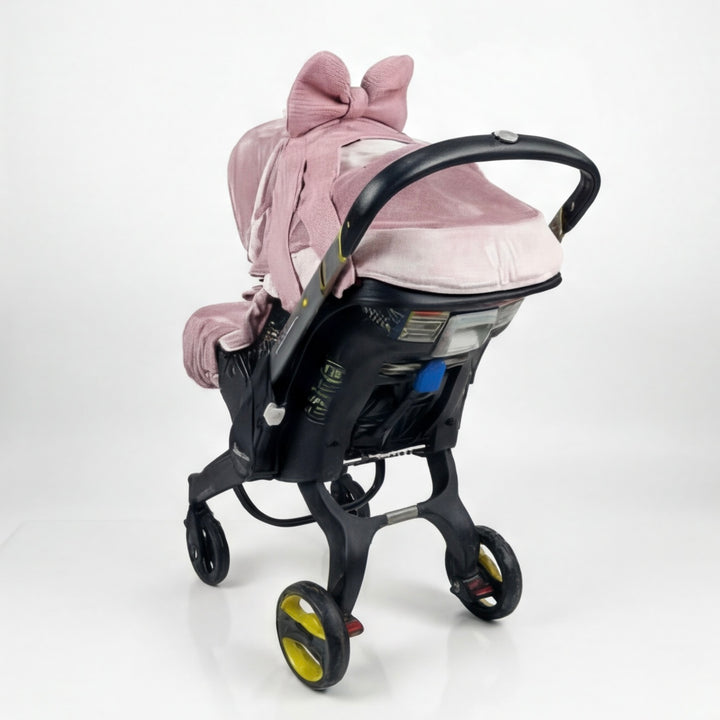 Rear three-quarter view of blush pink velvet Doona+ car seat cover set with large textured bow on canopy and matching footmuff bow, installed on Doona+ stroller mode, white studio background.