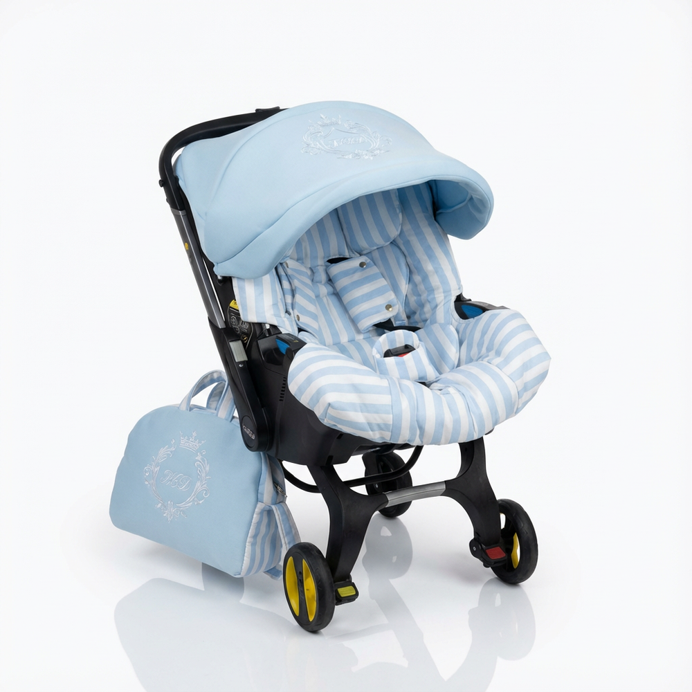 Front-facing view of an infant car seat on a black wheeled frame, featuring a light blue and white striped padded seat insert and matching light blue canopy with white embroidery, with a matching light blue diaper bag with white embroidery placed beside it on a light background.
