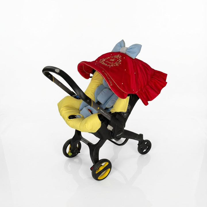 Red and light‑blue car seat cover set with satin quilting, gold “Victoria” embroidery, blue bows with pearl centers and yellow bow; white studio background.
