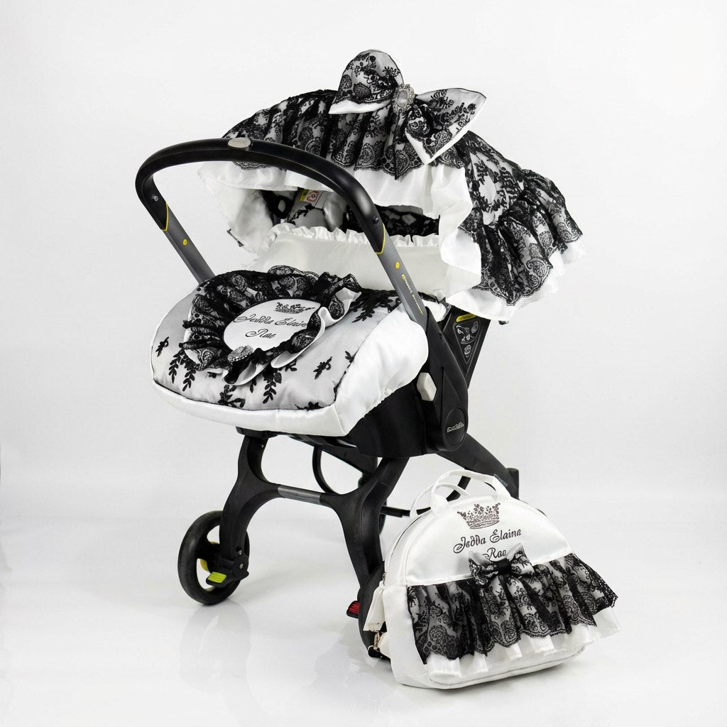 Front-view baby car seat cover set installed on a car seat, photographed on a white background. The design features a white base with black floral print accents, layered ruffled canopy with black lace trim, and a decorative bow on top. A matching padded footmuff with black lace details and a coordinated diaper bag are placed next to the car seat.