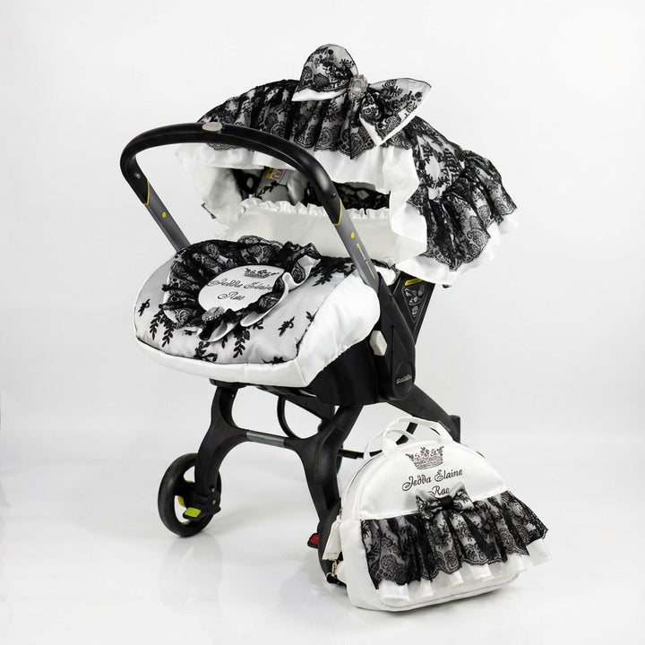 Front-view baby car seat cover set installed on a car seat, photographed on a white background. The design features a white base with black floral print accents, layered ruffled canopy with black lace trim, and a decorative bow on top. A matching padded footmuff with black lace details and a coordinated diaper bag are placed next to the car seat.