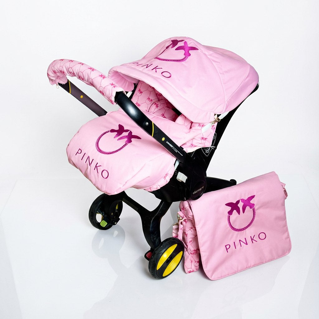 Pink baby car seat set installed on a car seat, featuring a soft pink canopy, padded footmuff, seat cover and handlebar cover with Pinko monogram embroidery, and a matching diaper bag. Coordinated pink patterned fabric details. Photographed on a clean white background.
