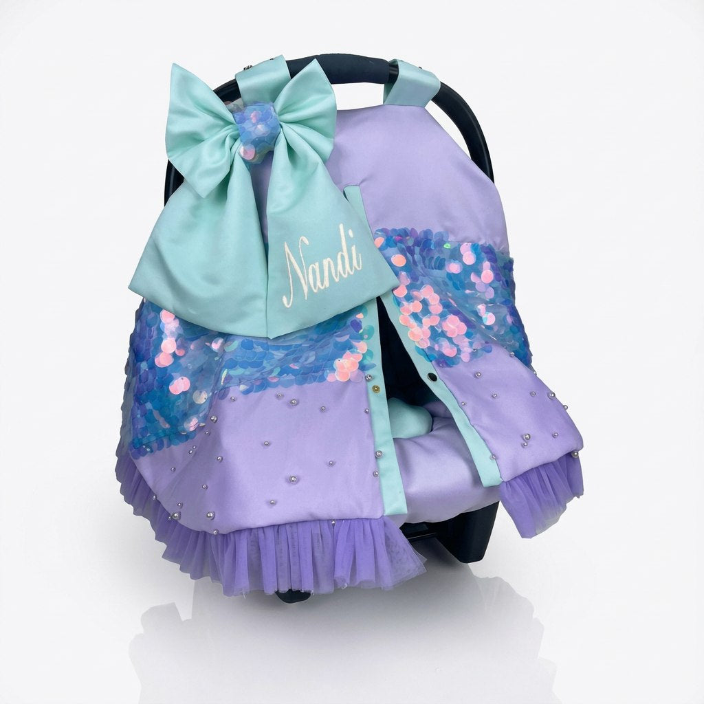 Front view of a baby car seat set installed on a car seat, featuring a lavender seat cover with mint green accents, decorative blue and pink sequin panels, small rhinestone embellishments, and a soft tulle ruffle along the bottom. The set includes a large mint bow with matching sequins and personalized name embroidery on the front. Displayed on a white background.