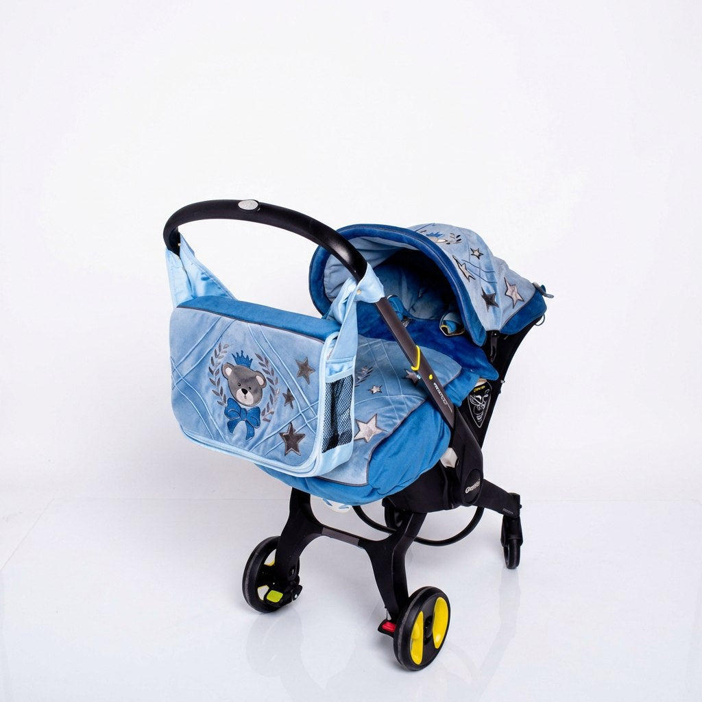 Side view of a Doona car seat on a light background, featuring a light blue quilted canopy and matching footmuff decorated with star patterns and an embroidered teddy bear emblem. A coordinated light blue bag with quilted panels and embroidered bear motif is attached to the handle.