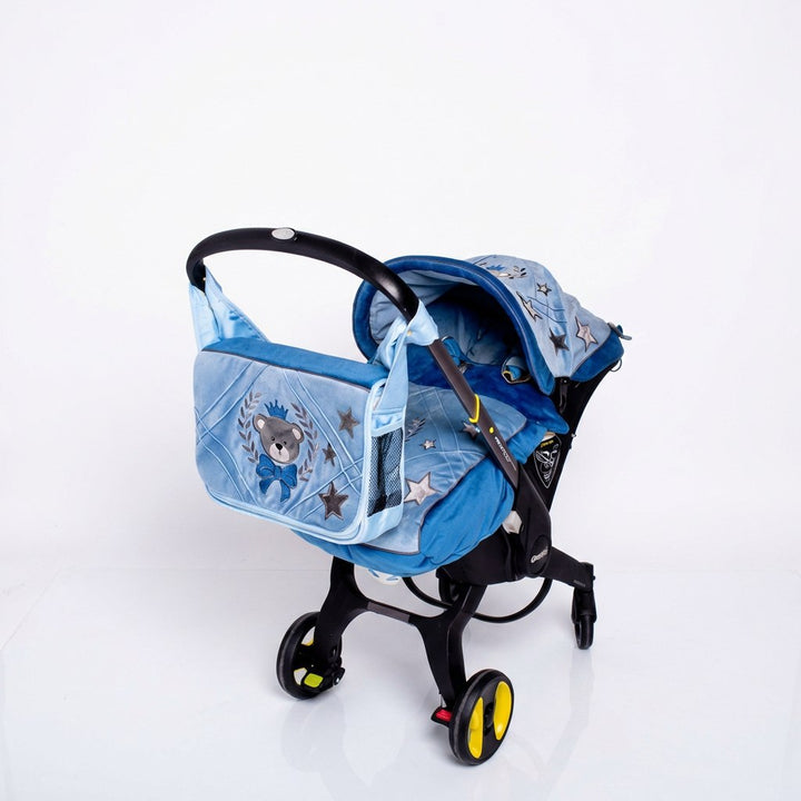 Side view of a Doona car seat on a light background, featuring a light blue quilted canopy and matching footmuff decorated with star patterns and an embroidered teddy bear emblem. A coordinated light blue bag with quilted panels and embroidered bear motif is attached to the handle.