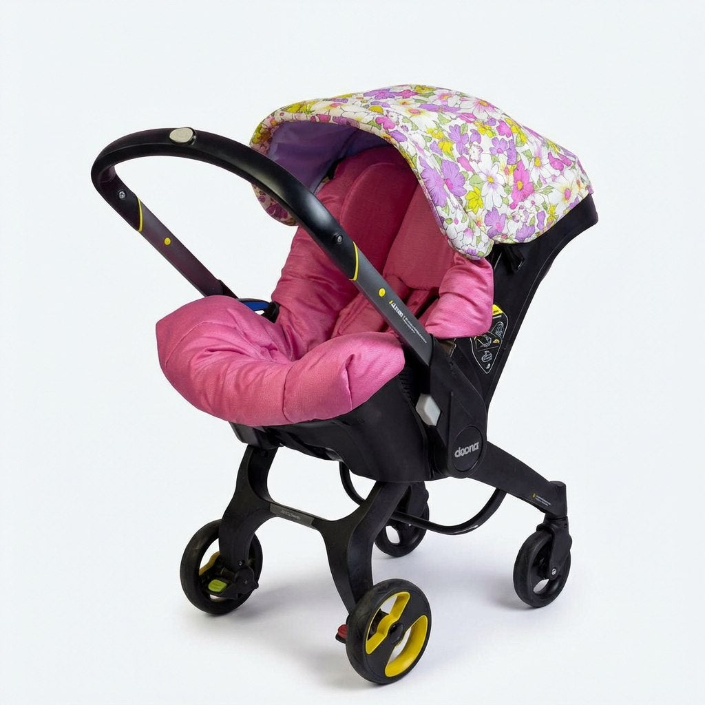 Infant car seat with deep pink fabric seat insert and floral canopy in pastel purple, yellow and white tones, installed on a black wheeled car seat frame on a light background.