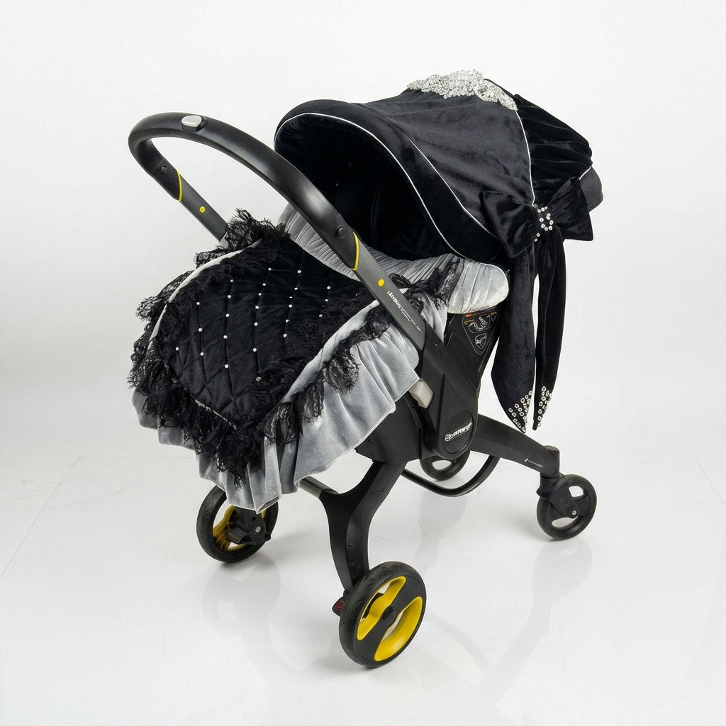 Black velour baby set installed on a car seat, featuring an elegant canopy with decorative hardware and crystal-embellished bow, a footmuff with soft gray ruffles, lace trim, and stone details, photographed on a clean white background.