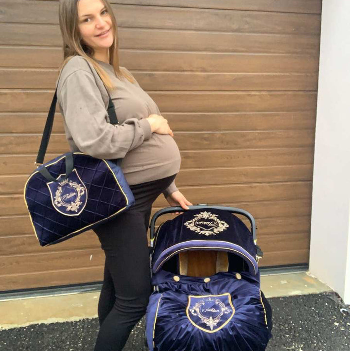 Expecting mother posing beside Royal Blue velvet Doona Plus car seat cover with gold piping, gold buttons and embroidered crest, styled with matching quilted blue and gold bag.