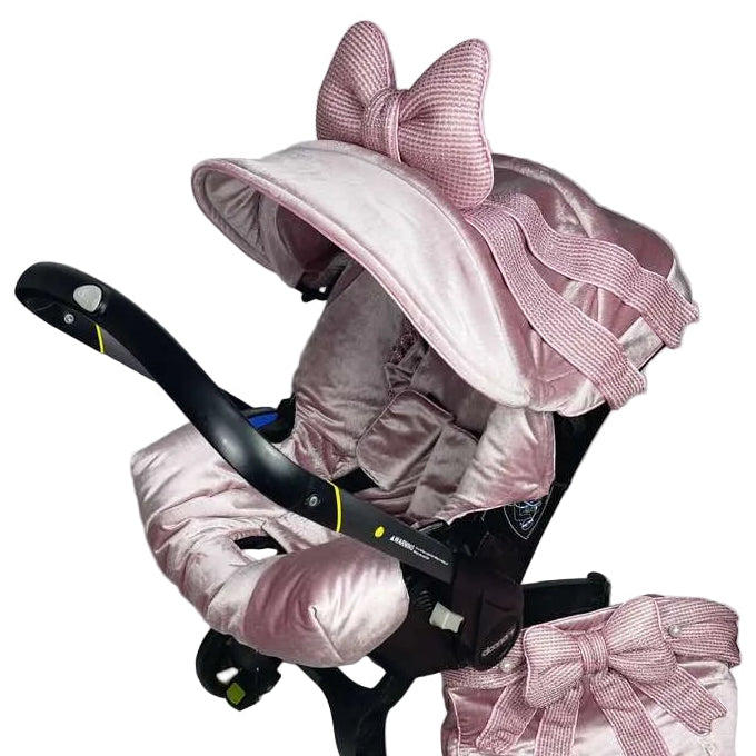 Rear three-quarter view of blush pink velvet Doona+ car seat cover set with large textured bow on canopy and matching footmuff bow, installed on Doona+ stroller mode, white studio background.
