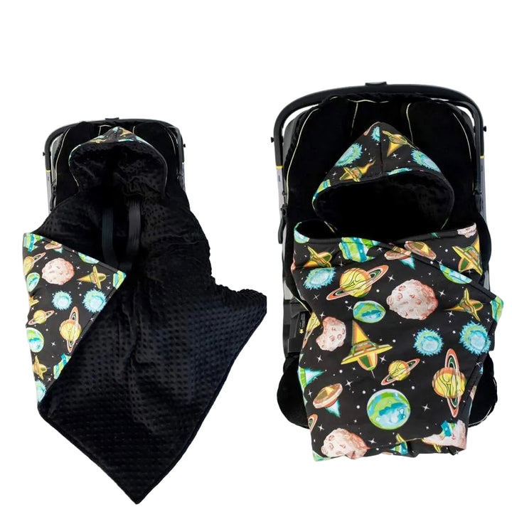 Front view of a hooded baby blanket that transforms into an envelope-style wrap, shown in two configurations. The left side shows the blanket partially opened with a black interior and space-themed outer fabric, while the right side shows the blanket fully wrapped into an envelope shape. The outer fabric features a colorful space print with planets, stars, and rockets, photographed on a light studio background.