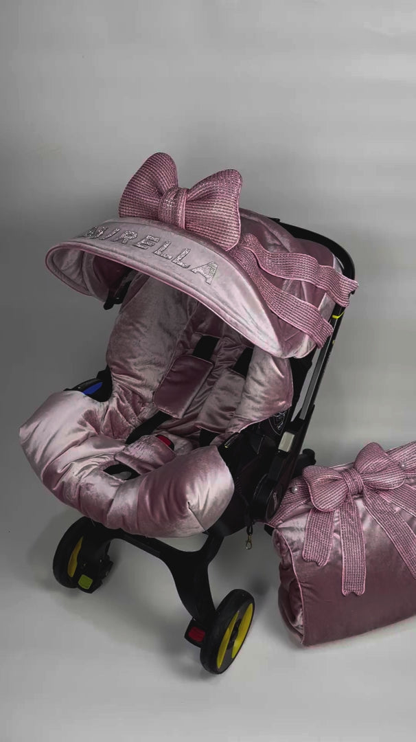 Video preview – side view of blush pink velvet Doona+ car seat cover set with large textured bow and long ribbon tails on canopy, matching bow footmuff and bag, shown in stroller mode on white studio background.